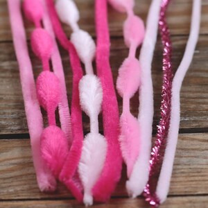 May include: A collection of pink, fuchsia, and white pipe cleaners and chenille stems. Some stems have fluffy, oval-shaped accents. A few stems are a sparkly, metallic pink. The items are arranged on a wooden surface.
