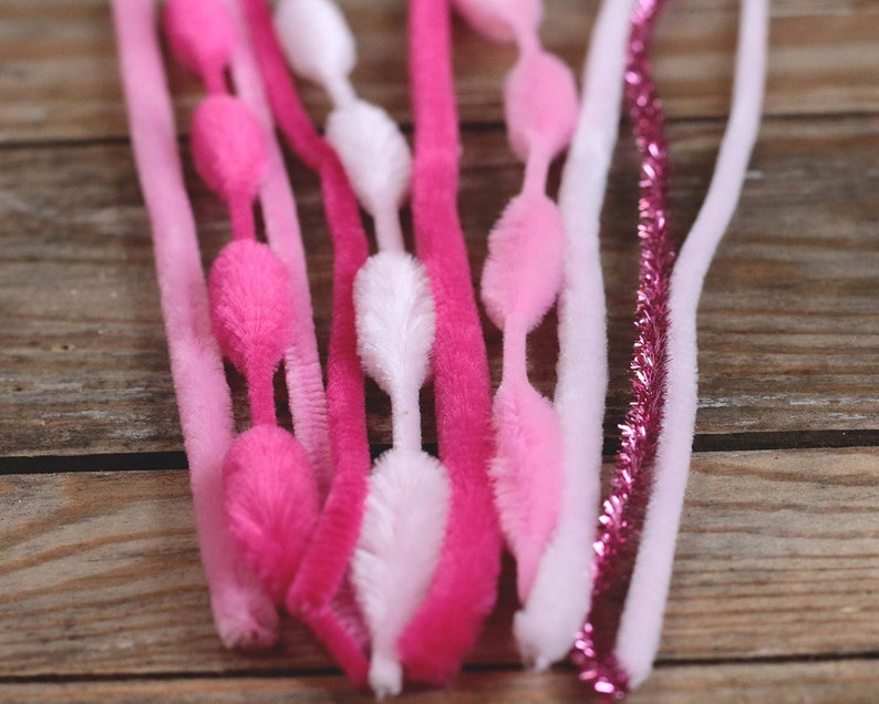 May include: A collection of pink, fuchsia, and white pipe cleaners and chenille stems. Some stems have fluffy, oval-shaped accents. A few stems are a sparkly, metallic pink. The items are arranged on a wooden surface.