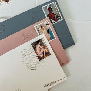 May include: A white envelope with a pink envelope and a blue envelope on top. The white envelope has a stamp with a nude woman on it and a stamp that says "Royal Postal Set - United Kingdom". The pink envelope has a stamp with a woman in a white dress on it and a stamp with a black woman on it. The blue envelope has a stamp with a woman in a white dress on it.