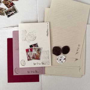May include: A set of stationery supplies with a vintage aesthetic. The set includes lined paper, envelopes, and decorative wax seals. The envelopes are pink and burgundy with a decorative stamp on each. The paper is cream colored with lines and a decorative border. The wax seals are brown and white with a floral design.
