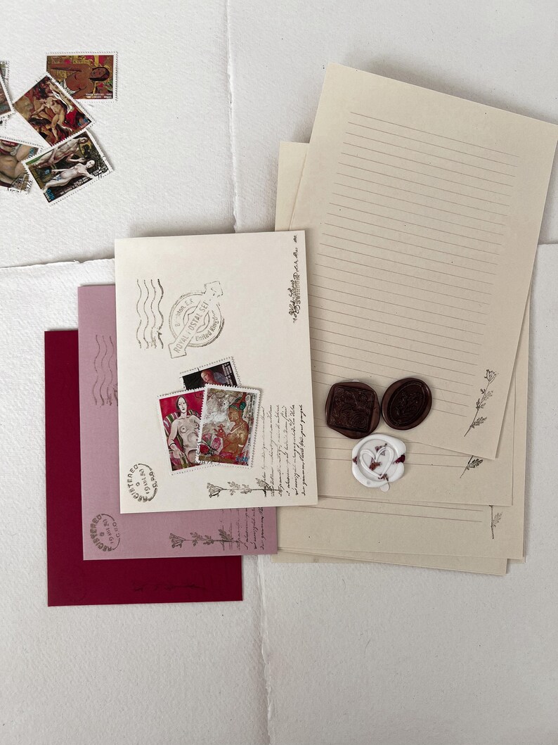May include: A set of stationery supplies with a vintage aesthetic. The set includes lined paper, envelopes, and decorative wax seals. The envelopes are pink and burgundy with a decorative stamp on each. The paper is cream colored with lines and a decorative border. The wax seals are brown and white with a floral design.