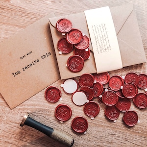 Peut inclure: Une enveloppe brune avec le texte "Open when you receive this" sur le devant. L'enveloppe est décorée de sceaux de cire rouge avec un motif floral.