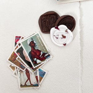 May include: A collection of vintage postage stamps featuring a variety of designs, including a woman with a crown and a portrait of a woman. The stamps are arranged in a pile on a white surface. There are also two wax seals, one brown and one white, with floral designs.