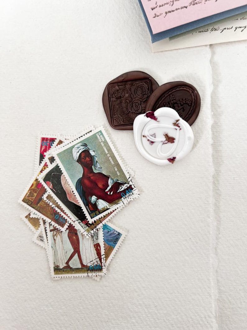 May include: A collection of vintage postage stamps featuring a variety of designs, including a woman with a crown and a portrait of a woman. The stamps are arranged in a pile on a white surface. There are also two wax seals, one brown and one white, with floral designs.