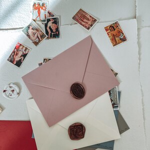 May include: Three envelopes with wax seals. The top envelope is pink, the middle envelope is white, and the bottom envelope is red. The envelopes are on a white surface with vintage postage stamps scattered around them.