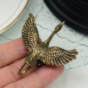 May include: A gold-toned metal crane figurine with outstretched wings. The crane is in flight with its head turned to the side. The figurine is detailed with textured feathers.