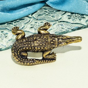 May include: A small, gold-colored metal figurine of a crocodile. The crocodile is posed with its head turned to the side and its mouth slightly open. The figurine is detailed with scales and textured skin.