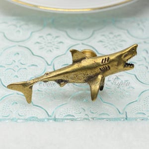 May include: A gold-toned metal shark fin cabinet knob or drawer pull. The shark is facing right with its mouth open.
