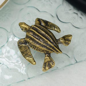 Op de afbeelding: Een klein, goudkleurig metalen beeldje van een lederschildpad. De schildpad is afgebeeld in een realistische houding met zijn flippers uitgespreid.