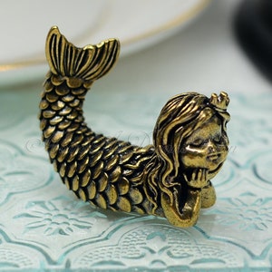 Könnte beinhalten: Eine goldfarbene Meerjungfrauenfigur mit einer Krone auf dem Kopf, die ihr Kinn auf ihre Hand legt. Sie hat einen langen, fließenden Schwanz mit Schuppen und sitzt auf einer hellblauen, gemusterten Oberfläche.