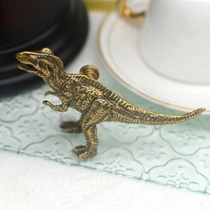 Puede incluir: Tirador de armario con forma de dinosaurio en latón antiguo detallado. El tirador es una miniatura de Tyrannosaurus Rex, con detalles intrincados en su cuerpo. El dinosaurio está de pie sobre sus patas traseras, con los brazos extendidos. Para armario o cajón.