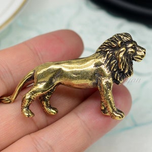 Può includere: Una piccola statuetta di leone in ottone dorato. Il leone è in piedi su tutte e quattro le zampe con la testa girata di lato. La statuetta è dettagliata con una criniera e una coda.