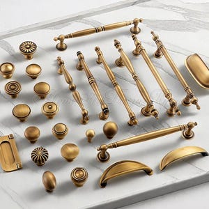 May include: An assortment of antique brass cabinet knobs and pulls in various shapes and sizes. The hardware includes round knobs, elongated pulls, and cup pulls, all with a warm, aged finish. The hardware is arranged on a white surface.