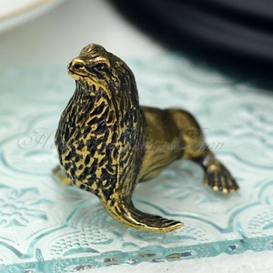 May include: A small, gold-coloured figurine of a seal. The seal is sitting on a glass surface with a decorative pattern.