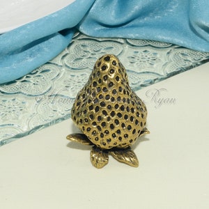 May include: A gold-coloured metal strawberry figurine with a detailed textured surface. The figurine is sitting on a glass surface with a decorative pattern.