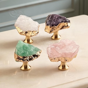 Puede incluir: Cuatro pomos para armarios con piedras de cristal en bruto e irregulares en blanco, morado oscuro, verde claro y rosa pálido. Cada piedra se asienta sobre una base de metal dorado pulido con un borde texturizado y orgánico.