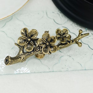 May include: Gold-toned metal cabinet knob featuring a design of a branch with three stylised cherry blossoms.