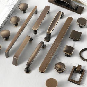 以下が含まれることがあります： ブロンズ調のキャビネット金具の品揃え。ノブ、プル、ハンドルが含まれています。丸いノブから長方形のプルやハンドルまで、さまざまな形とサイズがあり、すべてヴィンテージ感があります。明るい色の表面に配置されています。