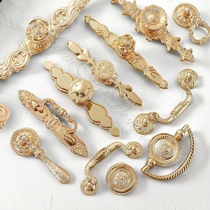 May include: An assortment of gold-coloured cabinet handles and knobs with intricate designs. The hardware features floral and geometric patterns, giving a vintage feel. Various sizes and styles are displayed on a white surface.