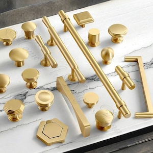 May include: A collection of gold-toned cabinet hardware, including knobs and pulls, arranged on a marble-patterned surface. The hardware features various shapes, such as round, square, and bar styles, offering a range of design options for furniture and cabinetry.