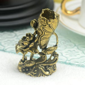 Puede incluir: Un soporte decorativo de metal dorado con un diseño de flor de loto. El soporte tiene una abertura en forma de jarrón en la parte superior y presenta dos peces a cada lado. El soporte está sobre una superficie blanca.