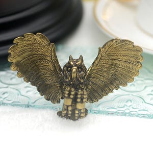 May include: A gold-colored owl figurine with outstretched wings. The owl is detailed with feathers and has a beaded scarf around its neck.