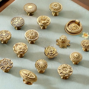 May include: A collection of antique brass cabinet knobs with various floral and geometric designs. The knobs are in different shapes and sizes, including round, oval, and flower-shaped, with intricate detailing. The knobs are displayed on a light blue surface.