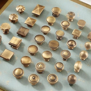 Op de afbeelding: Een verzameling antieke messing kastknoppen in diverse vormen en ontwerpen. De knoppen omvatten ronde, vierkante en facetstijlen, allemaal met een warme, verouderde afwerking. Ze zijn gerangschikt op een lichtblauwe, bedrukte ondergrond.