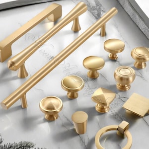 Puede incluir: Una colección de herrajes dorados para armarios, que incluye varios tiradores y pomos. El surtido presenta diferentes formas y tamaños, como tiradores de barra, pomos redondos y tiradores cuadrados. Los herrajes se exhiben sobre una superficie de mármol.