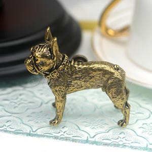 May include: A gold-toned metal figurine of a French Bulldog with a collar. The figurine is standing on a light blue surface with a decorative pattern.