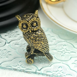 Op de afbeelding: Een gouden uilbeeldje dat op een tak zit. De uil heeft grote, ronde ogen en gedetailleerde veren.