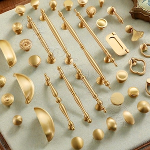 Può includere: Una collezione di ferramenta dorata per mobili, tra cui pomelli, maniglie e tiranti, disposti su una superficie chiara. La ferramenta presenta vari design, dai semplici pomelli rotondi alle maniglie ornate, offrendo una gamma di opzioni.