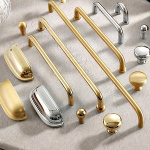 Op de afbeelding: Een assortiment gouden en zilveren kastbeslag, waaronder handgrepen en knoppen. De handgrepen zijn lang en gebogen, terwijl de knoppen rond zijn. Het beslag wordt gepresenteerd op een neutraal gekleurd oppervlak, dat de verscheidenheid aan ontwerpen en afwerkingen laat zien.