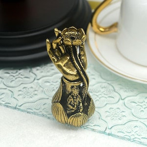 Peut inclure: Un brûle-encens en métal doré en forme de main tenant une fleur de lotus. La main tient une petite figurine d'une personne assise dans la fleur de lotus.
