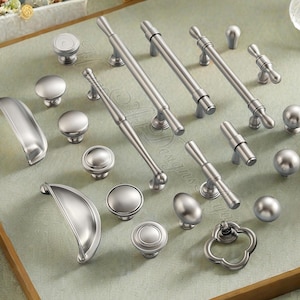 Puede incluir: Una colección de herrajes de gabinete plateados, que incluyen perillas, jaladeras y manijas. Los herrajes presentan varios diseños, desde perillas redondas simples hasta jaladeras de barra alargadas y manijas decorativas. Las piezas están dispuestas sobre una superficie verde claro.