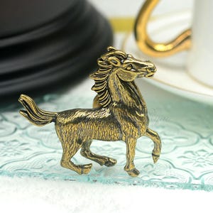 Könnte beinhalten: Eine detaillierte Pferdfigur aus Messing im antiken Stil. Das Pferd ist im Schritt dargestellt, mit aufwendigen Details in Mähne, Schweif und Körper. Die Figur hat eine warme, goldene Farbe, die auf eine gealterte oder Vintage-Ästhetik hindeutet.