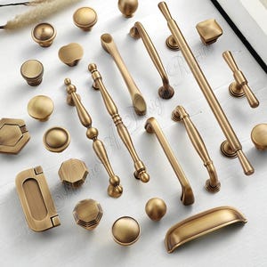 Op de afbeelding: Een assortiment antiek messing kastbeslag, waaronder knoppen en handgrepen in diverse vormen en maten. Het beslag heeft een warme, gouden tint en is gerangschikt op een wit oppervlak. De collectie omvat ronde, vierkante en langwerpige ontwerpen.