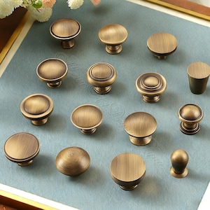 Op de afbeelding: Een verzameling antieke messing kastknoppen in diverse vormen en maten. De knoppen hebben een warme, verouderde afwerking, met platte, afgeronde en geribbelde oppervlakken. De hardware is gerangschikt op een lichtblauwe ondergrond, wat duidt op woonaccessoires.