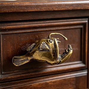 May include: A brass-coloured drawer pull shaped like an anglerfish, mounted on a dark wooden cabinet. The fish has detailed fins, an open mouth, and a curved lure. The cabinet has a recessed panel design.