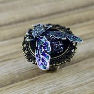 Może przedstawiać: Srebrny pierścionek z ozdobnym, bogato zdobionym wzorem przedstawiającym zielono-fioletową pszczołę wysadzaną klejnotami.