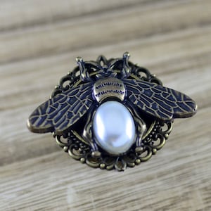 Könnte beinhalten: Eine goldfarbene Bienenbrosche mit einem weißen Perlenmittelpunkt. Die Biene ist mit filigranen Flügeln und einem filigranen Design verziert.