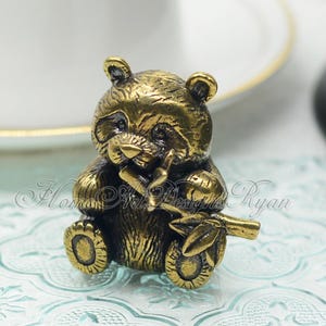 May include: A small, gold-coloured metal figurine of a panda bear sitting and holding a piece of bamboo in its paws.