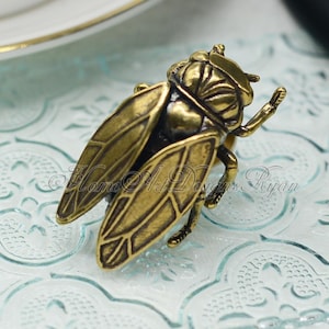 May include: A gold-toned cicada insect figurine with detailed wings and legs. The figurine is sitting on a light blue patterned surface.