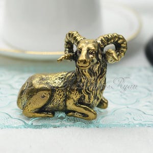 May include: A detailed, gold-coloured ram figurine in a seated position. The ram has large, curved horns and is resting on a light blue, patterned surface. The figurine is likely made of metal and has intricate detailing.