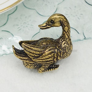 以下が含まれることがあります： 金色をした小さな金属製の鴨の置物。鴨は頭を横に傾け、羽を広げて座っている。