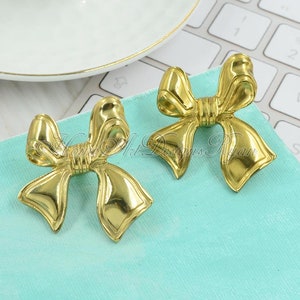 Puede incluir: Dos pendientes de color dorado en forma de lazo. Los pendientes están sobre una superficie azul claro.