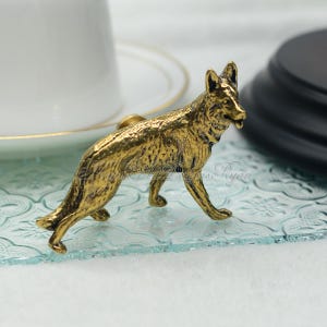 May include: A gold-colored metal German Shepherd dog figurine. The figurine is standing on a light blue glass surface.