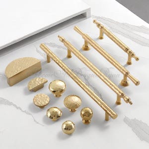 May include: A collection of gold cabinet hardware, including handles and knobs, arranged on a white surface. The hardware features a hammered texture and various shapes, including half-moon and round designs. The image showcases a variety of sizes and styles.