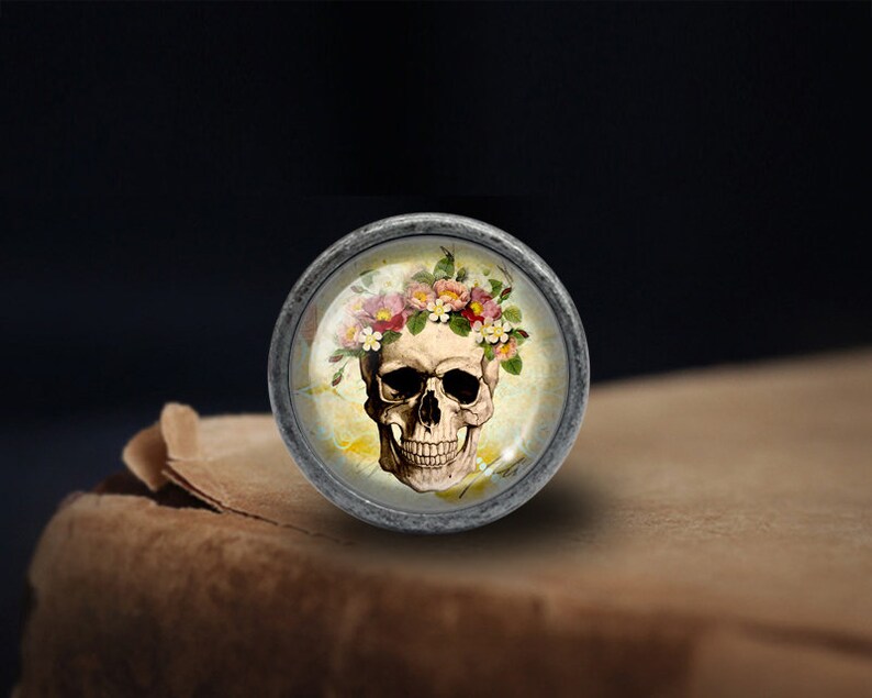 K&ouml;nnte beinhalten: Ein silberner Knopf mit einem Sch&auml;del, der eine Blumenkrone tr&auml;gt. Der Sch&auml;del ist wei&szlig; mit schwarzen Augenh&ouml;hlen und einem schwarzen Mund. Die Blumen sind rosa, wei&szlig; und gelb.