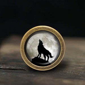 Może przedstawiać: Okrągły, mosiężny, ozdobny gałka z wkładką szklaną przedstawiającą czarną sylwetkę wilka wyjącego do pełnego księżyca.